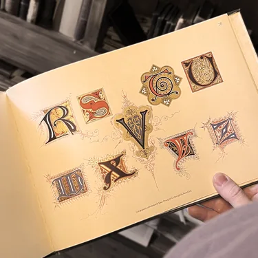 A hand holding an antique book of illuminated initial letters in front of library shelves